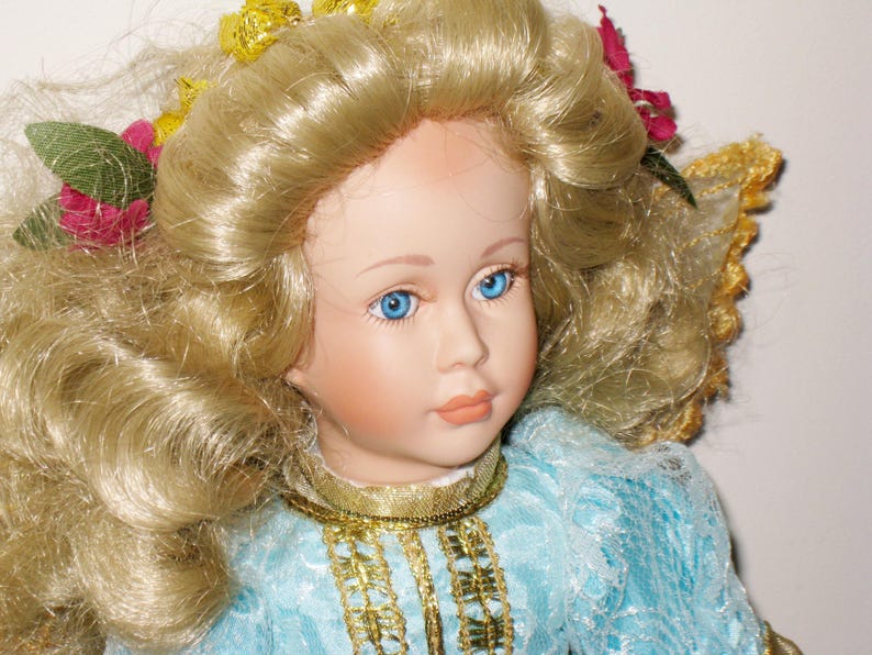 May include: A porcelain doll with blonde, curly hair and blue eyes. The doll wears a light blue dress with gold trim and a gold necklace. Floral accents are in the doll's hair.