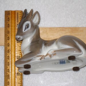 May include: A porcelain figurine of a fawn lying down. The fawn is painted in shades of brown and white, with a blue and white company name on the underside. The figurine is resting on a wooden surface.