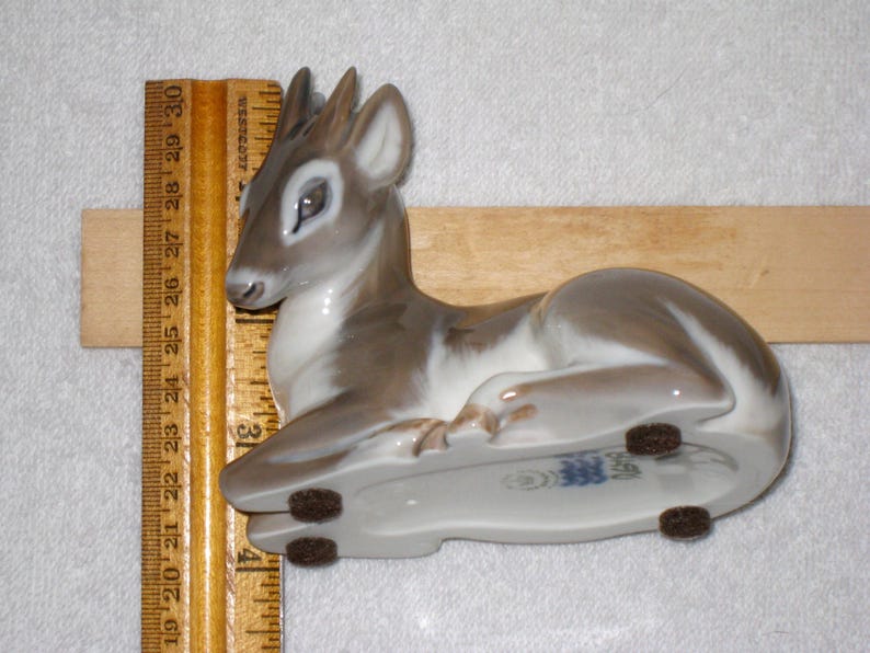 May include: A porcelain figurine of a fawn lying down. The fawn is painted in shades of brown and white, with a blue and white company name on the underside. The figurine is resting on a wooden surface.