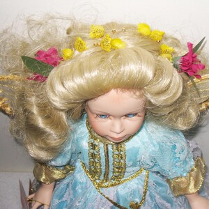 May include: A porcelain doll with blonde hair, blue eyes, and a light blue dress with gold trim. The doll has a floral headpiece with yellow and pink accents and gold-trimmed wings.