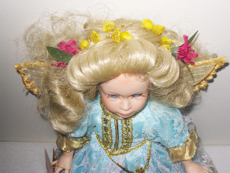 May include: A porcelain doll with blonde hair, blue eyes, and a light blue dress with gold trim. The doll has a floral headpiece with yellow and pink accents and gold-trimmed wings.