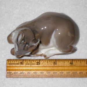 May include: A porcelain figurine of a fawn lying down with its head resting on its front legs. The fawn is brown and white with a glazed finish. The figurine is approximately 4 1/2 inches long and is sitting on a wooden ruler with markings for inches.