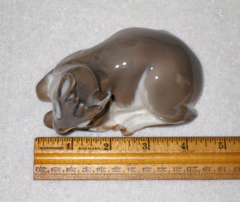 May include: A porcelain figurine of a fawn lying down with its head resting on its front legs. The fawn is brown and white with a glazed finish. The figurine is approximately 4 1/2 inches long and is sitting on a wooden ruler with markings for inches.