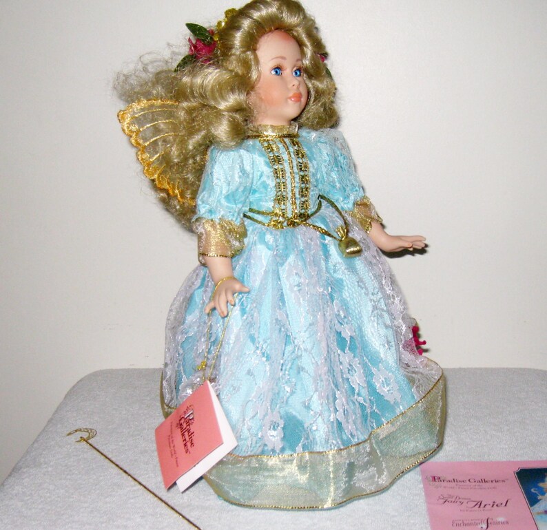 May include: A porcelain fairy doll with blonde curly hair, blue eyes, and gold-trimmed wings. The doll wears a light blue dress with white lace overlay and gold accents. A small tag hangs from the doll's hand. A gold wand rests on the surface.