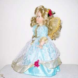 May include: A porcelain doll with blonde curly hair and blue eyes. The doll wears a light blue dress with gold trim and a white lace overlay. A red flower adorns the doll's hair. The doll has gold wings.