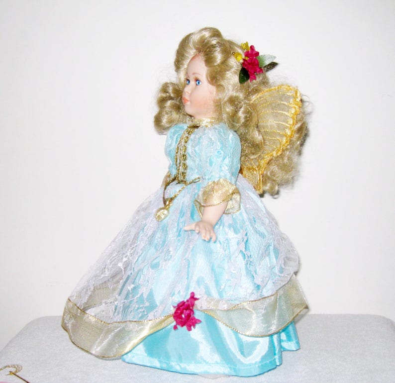 May include: A porcelain doll with blonde curly hair and blue eyes. The doll wears a light blue dress with gold trim and a white lace overlay. A red flower adorns the doll's hair. The doll has gold wings.
