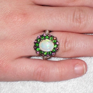 May include: A silver ring featuring a large oval opal gemstone surrounded by a halo of green and pink gemstones. The ring is set on a hand, showcasing the intricate design and vibrant colors of the jewelry.