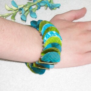 Könnte beinhalten: Ein farbenfrohes Armband mit schillernden, halbkreisförmigen Perlen in Türkis-, Blau- und Grüntönen. Das Armband wird am Handgelenk getragen und zeigt seine leuchtenden Farben und sein einzigartiges Design. Die Perlen sind durch ein flexibles Band verbunden.