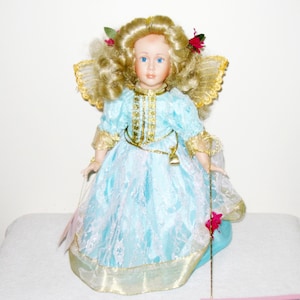 May include: A porcelain doll with blonde curly hair, blue eyes, and a light blue dress with gold trim. The doll has gold wings and holds a gold staff. Red flowers and green leaves adorn the doll's hair. The doll is a decorative collectible.
