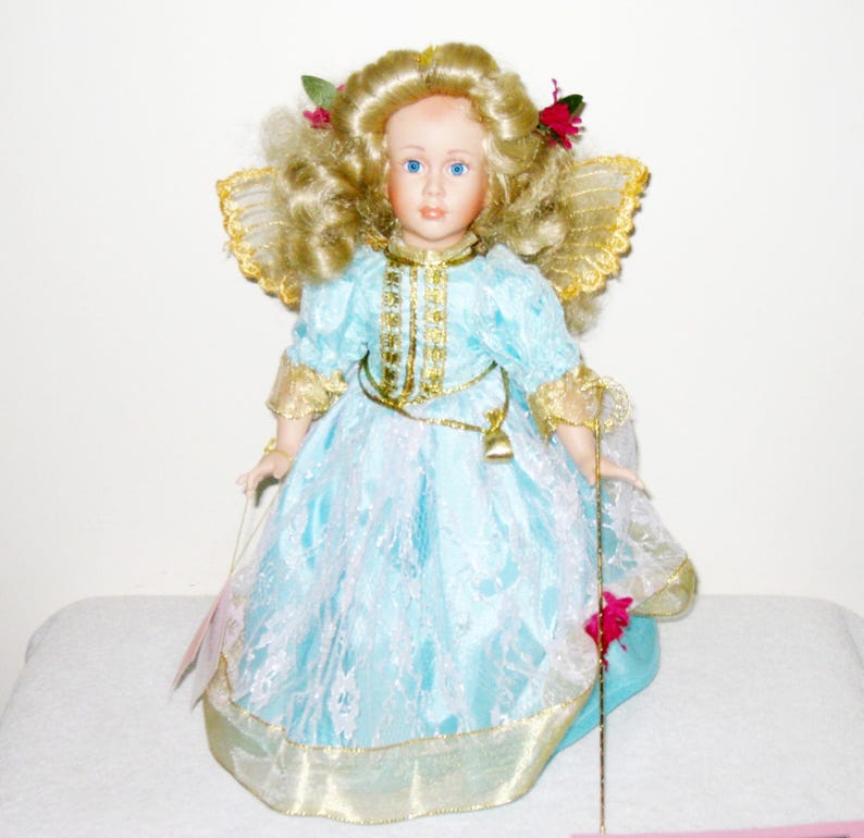 May include: A porcelain doll with blonde curly hair, blue eyes, and a light blue dress with gold trim. The doll has gold wings and holds a gold staff. Red flowers and green leaves adorn the doll's hair. The doll is a decorative collectible.