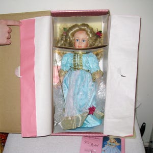 May include: A porcelain doll with blonde curly hair and blue eyes, wearing a light blue dress with gold trim, presented in a decorative box. The doll has a delicate face and is adorned with a red rose. The box is lined with pink paper.