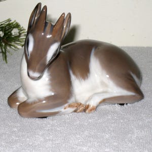 May include: A porcelain figurine of a fawn lying down. The fawn is brown and white with a detailed face and antlers.
