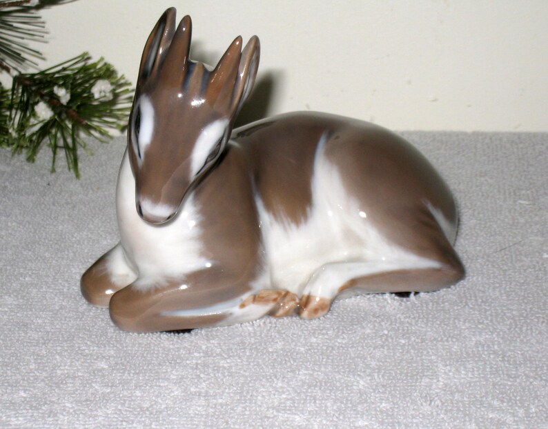 May include: A porcelain figurine of a fawn lying down. The fawn is brown and white with a detailed face and antlers.