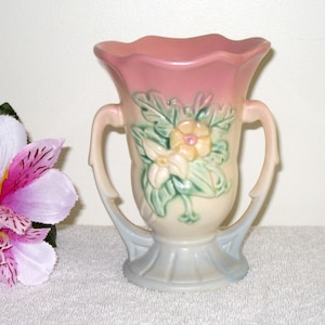 May include: A vintage ceramic vase with a pink and cream gradient. The vase features two handles and a floral design with yellow and white flowers and green leaves. The base is a light blue color.