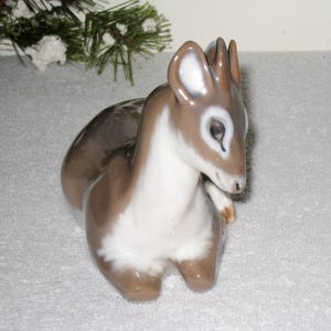 May include: A porcelain figurine of a fawn lying down with its head turned to the side. The fawn is brown and white with a detailed face and large ears.