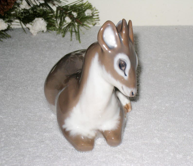 May include: A porcelain figurine of a fawn lying down with its head turned to the side. The fawn is brown and white with a detailed face and large ears.