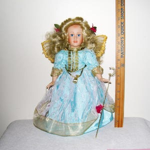 May include: A porcelain doll with blonde curly hair, blue eyes, and a light blue dress with gold trim. The doll has a gold necklace and holds a silver staff. The doll is approximately 12 inches tall.