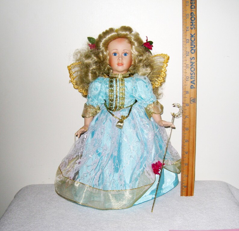 May include: A porcelain doll with blonde curly hair, blue eyes, and a light blue dress with gold trim. The doll has a gold necklace and holds a silver staff. The doll is approximately 12 inches tall.