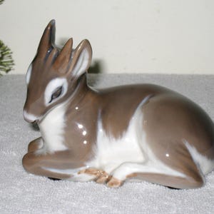 May include: A porcelain figurine of a fawn lying down. The fawn is brown and white with a glossy finish.