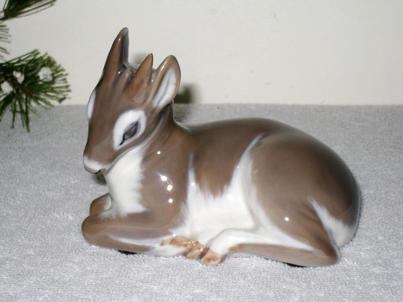 May include: A porcelain figurine of a fawn lying down. The fawn is brown and white with a glossy finish.