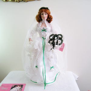May include: A porcelain doll in a white wedding dress with green accents, holding a bouquet. The doll has red hair and a veil. The dress features lace detailing and a green ribbon. A tag is attached to the bouquet.