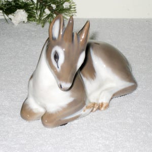 May include: A porcelain figurine of a fawn with brown and white markings. The fawn is sitting with its head down, its eyes open. The figurine is made by Royal Copenhagen and is part of their animal collection.