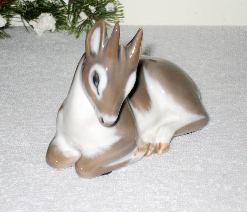 May include: A porcelain figurine of a fawn with brown and white markings. The fawn is sitting with its head down, its eyes open. The figurine is made by Royal Copenhagen and is part of their animal collection.
