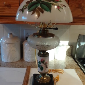 May include: A white glass lamp with a floral design on the shade and base. The lamp has a gold trim and a black base. The lamp is on a white surface.
