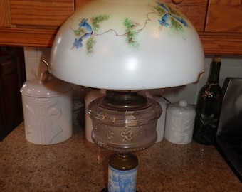 May include: A vintage oil lamp with a white glass shade featuring a floral design with blue flowers. The lamp has a clear glass base with a decorative pattern and a blue and white porcelain base.