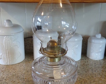 May include: A vintage glass oil lamp with a clear glass chimney and a cut glass base. The lamp is sitting on a black base and a white crocheted doily.