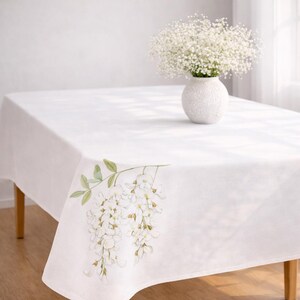 May include: A white rectangular tablecloth with a floral design in the corner. The design features white flowers and green leaves. A white vase with white flowers sits on the table.
