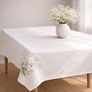 May include: A white rectangular tablecloth with a floral design in the corner. A white vase with white flowers sits on the table. The table has wooden legs.