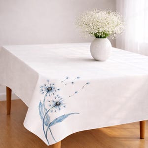 May include: A white rectangular tablecloth with a blue dandelion design on the corner. A white vase with white flowers sits on the table. The table has wooden legs.