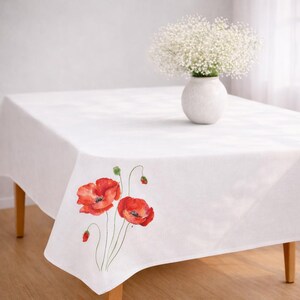 May include: White rectangular tablecloth with a watercolor design of red poppies and green buds. A white vase with white flowers sits on the table. The table has wooden legs.