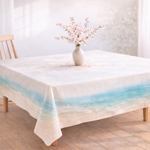 May include: Square tablecloth with a beach scene design. The tablecloth features a light blue and white ocean wave pattern. A white vase with pink flowers sits on the table. The table is made of wood.