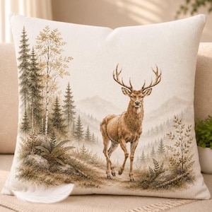 May include: A square throw pillow featuring a printed illustration of a brown deer standing in a forest landscape. The scene includes evergreen trees, birch trees, ferns, and rocks against a backdrop of mountains in muted earth tones.