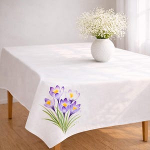 May include: A white rectangular tablecloth with a floral design featuring purple and yellow crocus flowers. A white vase with white flowers sits on the table. The tablecloth is on a wooden table.