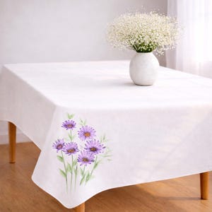 May include: A white tablecloth with a floral design featuring purple flowers and green stems. A white vase with a bouquet of white flowers sits on the table. The table has wooden legs.
