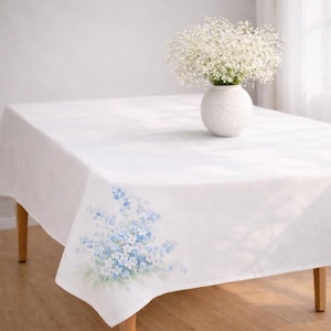 May include: A white rectangular tablecloth with a floral design in the corner. The floral design features blue and white flowers. A white vase with white flowers sits on the table.