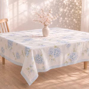 May include: A square tablecloth with a floral hydrangea pattern in shades of blue and green on a white background. The tablecloth is on a wooden table, and a white vase with pink flowers sits in the center. The tablecloth has a light blue border.