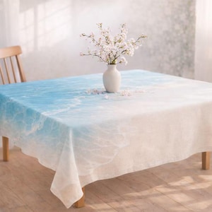 May include: A square tablecloth with a blue and white ocean wave design. A white vase with white flowers sits on the tablecloth. The tablecloth is on a wooden table with a wooden chair in the background.