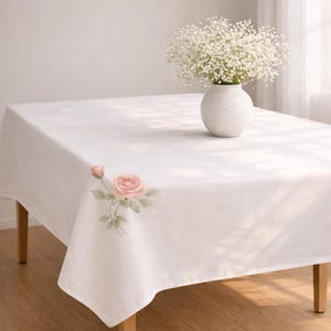 May include: A white rectangular tablecloth with a watercolor rose design in the corner. A white vase with a bouquet of white flowers sits on the table. The table has wooden legs.