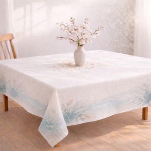 May include: A square white tablecloth with a blue and green watercolor design along the edges. A white vase with pink flowers sits in the center of the table. The table is made of wood and has a light brown color.