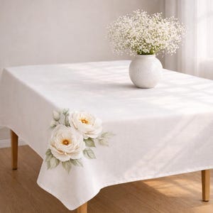 May include: A white rectangular tablecloth with a floral design featuring two large white roses with yellow centers and green leaves. A white vase with white flowers sits on the table. The table has wooden legs.