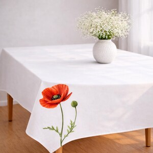 May include: A white rectangular tablecloth with a vibrant red poppy flower and bud design. A white vase with white flowers sits on the table. The tablecloth is draped over a wooden table.
