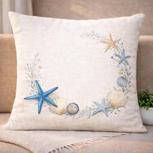 May include: A square, off-white pillow with a coastal design. The pillow features a wreath of seashells, starfish, and sprigs of greenery. The starfish are blue and tan. The pillow is on a neutral-colored sofa.