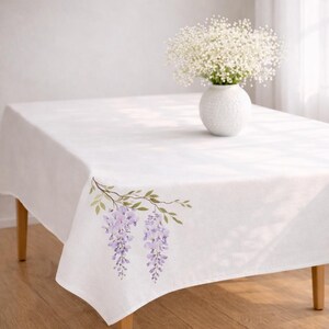 May include: White rectangular tablecloth with a floral design featuring lavender wisteria blossoms and green leaves. A white vase with white flowers sits on the table. The table has wooden legs.