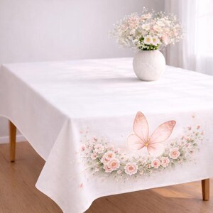 May include: A white tablecloth featuring a printed design of a large, soft pink butterfly centered above a curved arrangement of light pink roses and green foliage. A white vase filled with a bouquet of white and pink flowers sits on top.