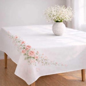 May include: A white tablecloth with a floral rose design in shades of pink and green along one edge. A white vase with a textured surface holds a bouquet of white flowers. The tablecloth is draped over a wooden table.
