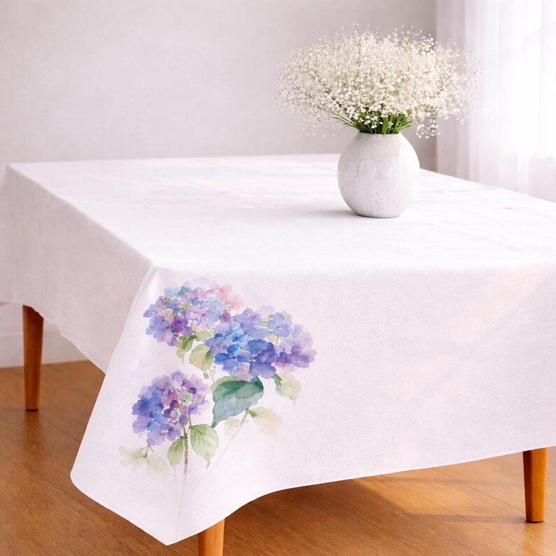May include: White rectangular tablecloth with a watercolour floral design of blue and purple hydrangeas. A white vase with a bouquet of white flowers sits on the table. The table has wooden legs.